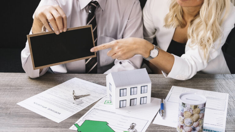 Rencontre entre un conseiller immobilier et une cliente dans une agence immobilière à Mulhouse, avec contrats, maquette de maison et clés sur la table.